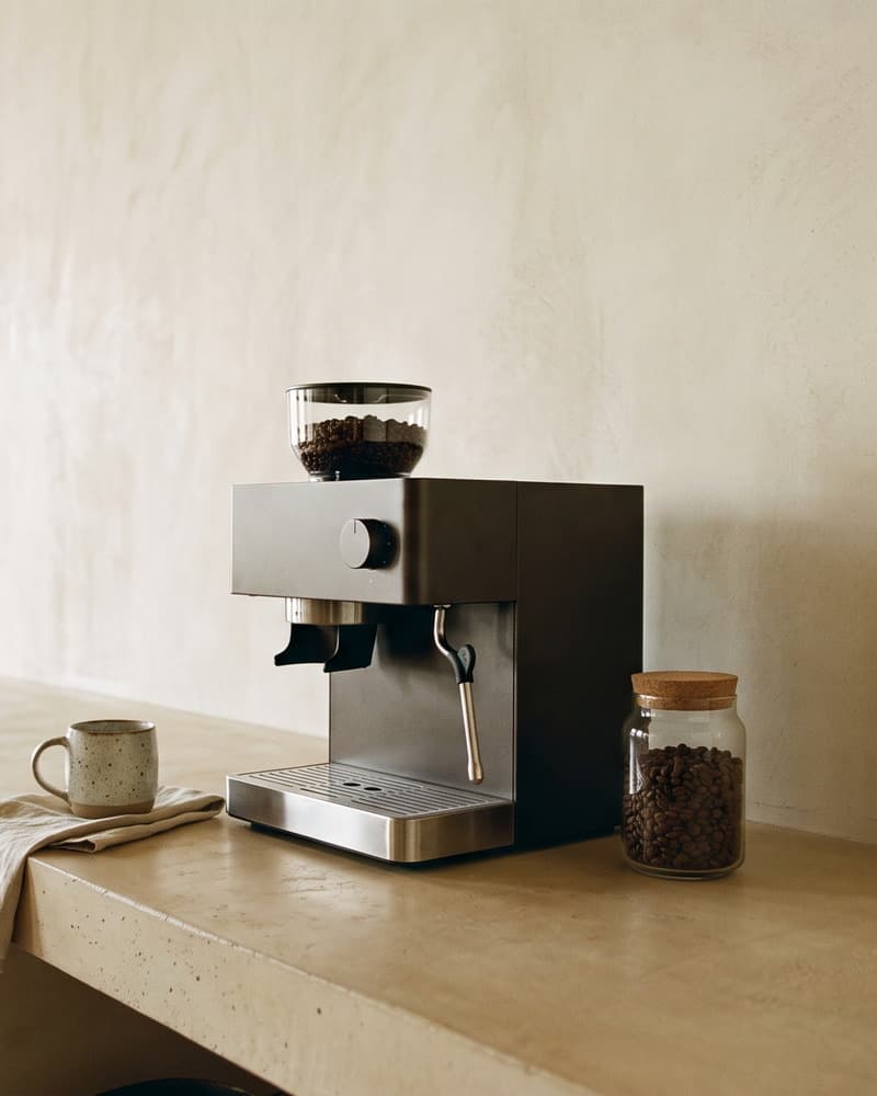 A long open counter of pale concrete along a bare plastered wall with nothing mounted above it: a plain machine with a bean hopper on top, a mug, a folded cloth and a jar of beans, open shelves beneath the counter, a window at the far end
