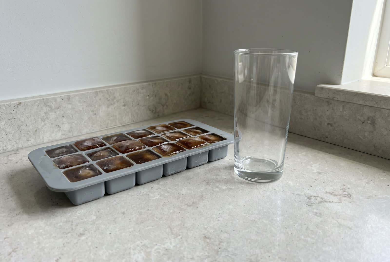 A silicone tray of frozen dark coffee cubes sits beside an empty tall glass on a stone counter.
