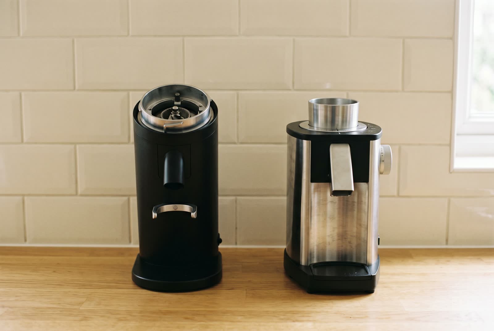 Two coffee grinders with their hoppers removed, one tall and narrow, one squat and wide, on a light oak counter.