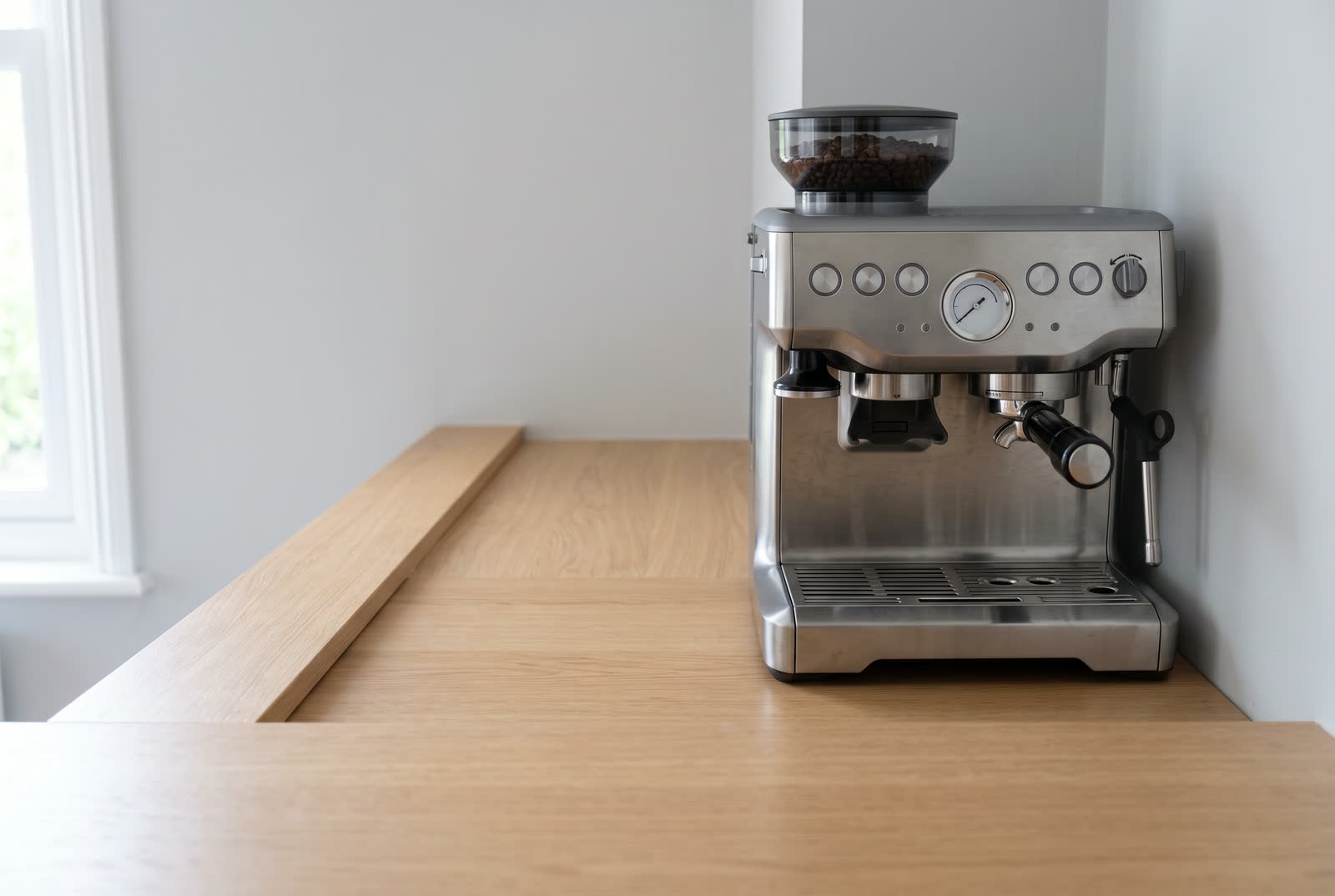 A narrow light oak counter run seen end on in perspective, only a little deeper than the machine itself, a brushed steel espresso machine backed up against the side wall that closes the run off with a hand's width of bare counter in front of it and the whole near half empty.