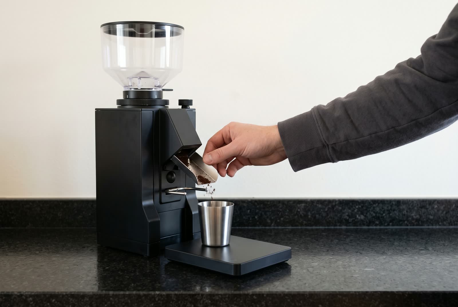 A hand steadies a grinder's open chute over a steel cup on a scale, loose grounds falling in.