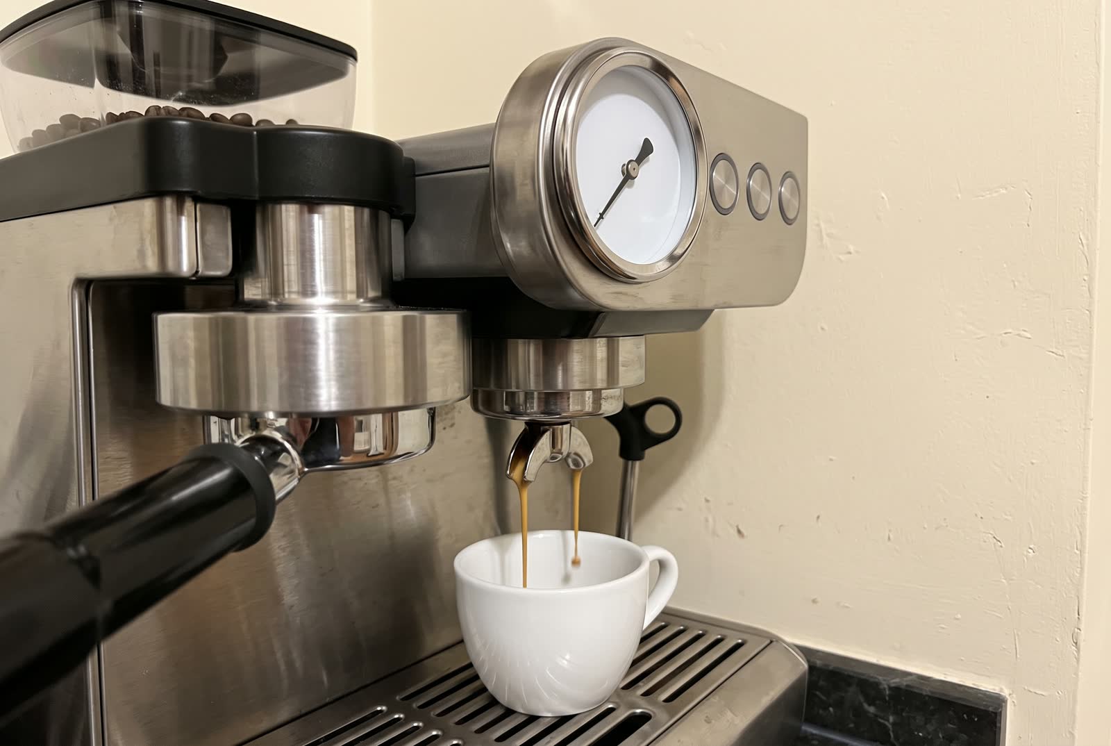 A close view of the working front of an espresso machine, the chrome group head with a loaded portafilter filling the lower left and the round pressure gauge the upper right, a thick dark stream running into a white cup below, the gauge face carrying one needle and one arc and no numerals.