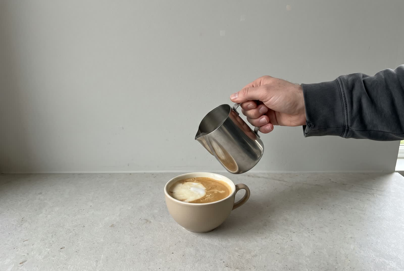 A jug pours over a cup of coffee, its surface showing a failed pour, a pale blob off to one side.