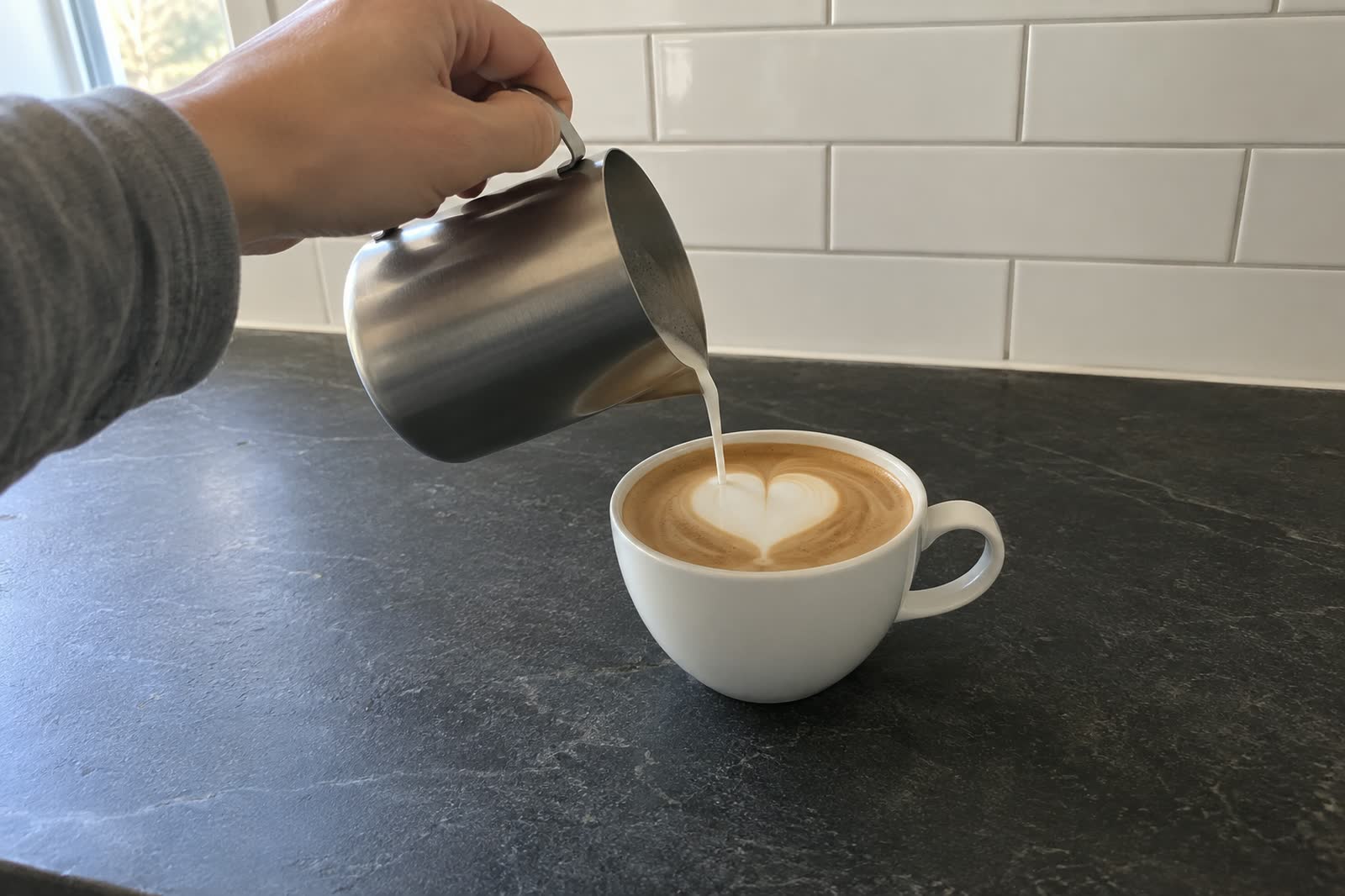 A hand in a grey sleeve tips a plain steel milk jug over a white cup on a dark soapstone counter, the stream of milk finishing a single heart in the foam, white subway tile behind.