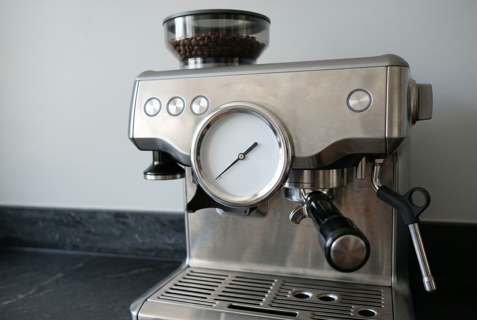 A close macro view of the round analogue pressure gauge on the front of a brushed steel espresso machine, its plain dial carrying one needle and one unbroken arc and no numerals, marks or lettering of any kind, the group head and portafilter handle soft behind it.