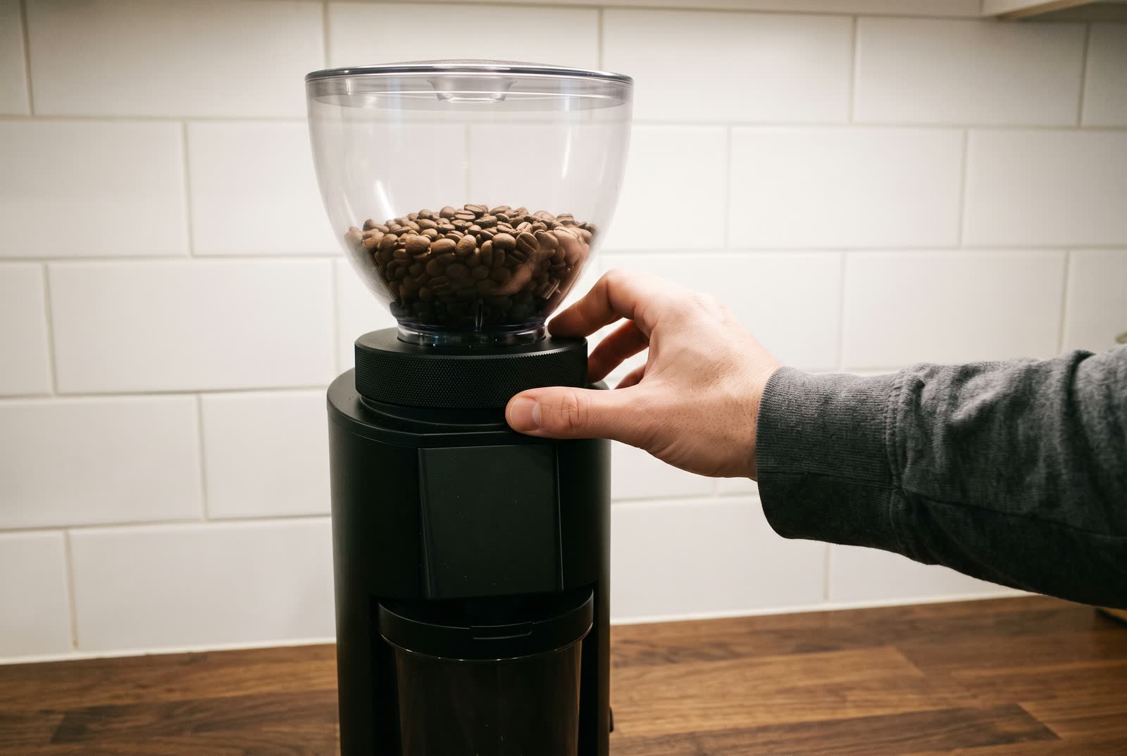 A hand turns the plain adjustment collar on a coffee grinder, its bean hopper full above.