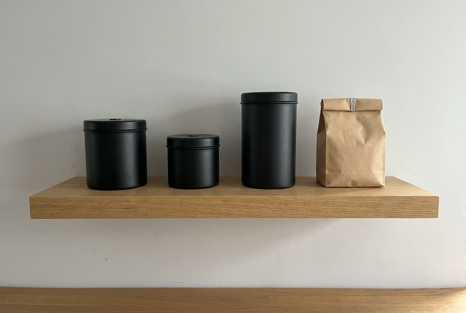 Three plain black canisters of three heights and a folded kraft coffee bag on a light oak shelf.